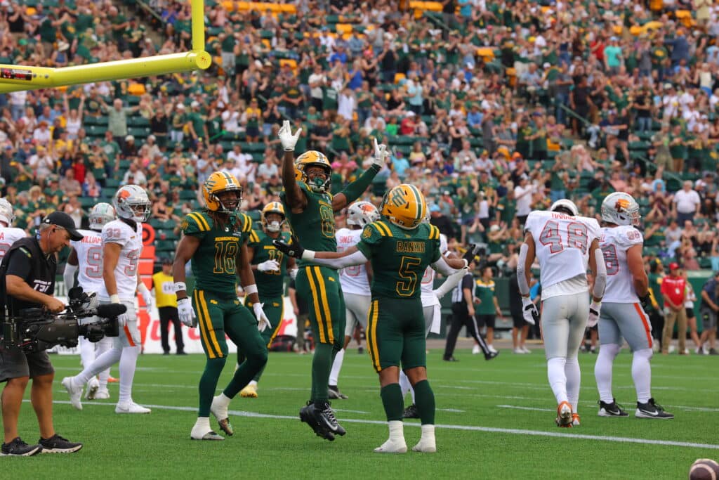 Edmonton Elks players in green and yellow uniforms celebrate on the field while the B.C. Lions in white uniforms stand nearby, with a large crowd in the stands behind them.