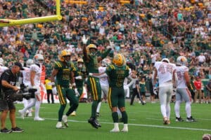 Edmonton Elks players in green and yellow uniforms celebrate on the field while the B.C. Lions in white uniforms stand nearby, with a large crowd in the stands behind them.